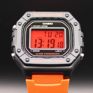 May include: A digital Casio watch with a black and gray case and an orange band. The watch face displays the date and time in red digits. The watch has the word "ILLUMINATOR" at the top and "WATER 50M RESIST" at the bottom.