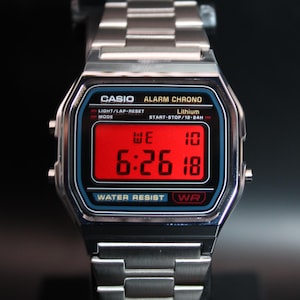 May include: A silver Casio digital watch with a rectangular face displaying the time 6:26:18. The watch face has a red digital display with the day and date. The watch band is made of silver metal links. The watch is labeled "CASIO ALARM CHRONO".