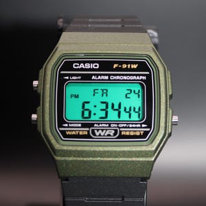 May include: A digital Casio F-91W watch with a green case and black strap. The watch face displays the time, day, and date in green digits. The watch has the Casio logo and model number printed on the face.