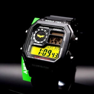 May include: Black Casio digital watch with a yellow display showing the time. The watch features a Batman logo and the text "WORLD TIME" and "ILLUMINATOR". The watch is on a black background.