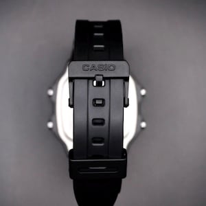 May include: A black Casio digital watch with a black strap. The watch strap is fastened with a black buckle that has the brand name "CASIO" printed on it. The watch face is silver and has a digital display.