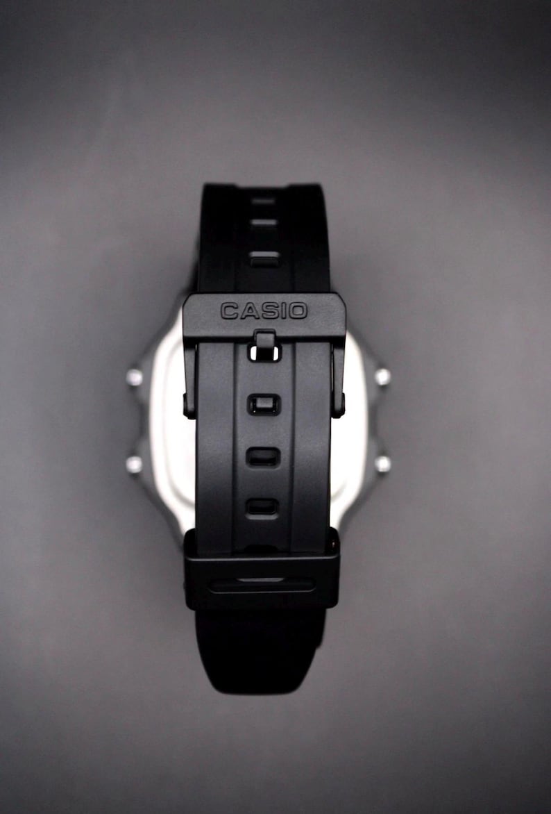 May include: A black Casio digital watch with a black strap. The watch strap is fastened with a black buckle that has the brand name "CASIO" printed on it. The watch face is silver and has a digital display.