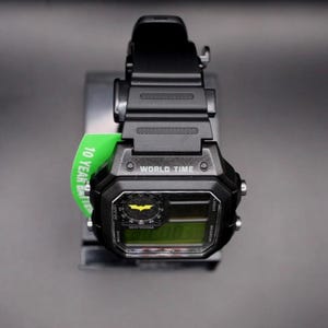 May include: Black digital watch featuring a Batman logo on the face. The watch has a black strap and a green tag that reads "10 Year Battery". The watch face displays "WORLD TIME".