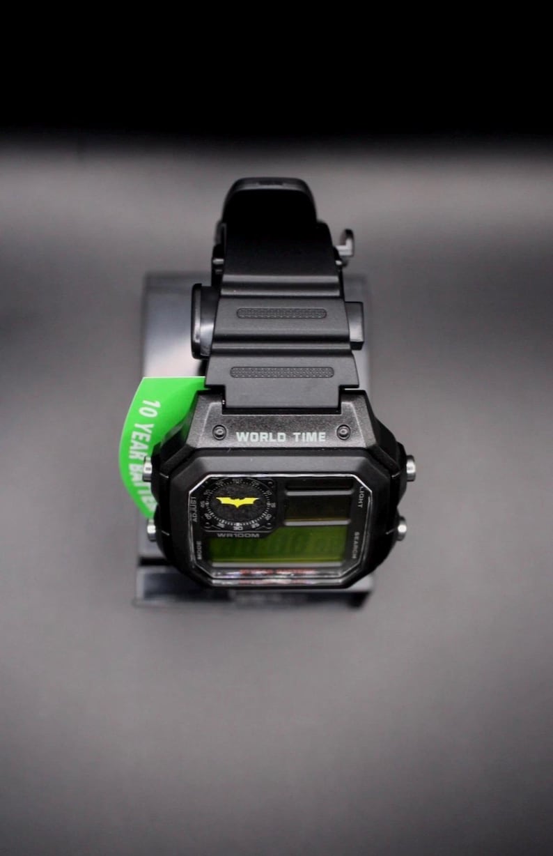 May include: Black digital watch featuring a Batman logo on the face. The watch has a black strap and a green tag that reads "10 Year Battery". The watch face displays "WORLD TIME".
