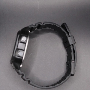 May include: A black digital wristwatch with a black rubber strap. The watch face has three buttons and a digital display. The strap is textured and has a buckle closure. The watch is designed for everyday wear.