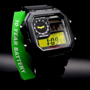 May include: Black Casio digital watch with a yellow display showing the time. The watch features a Batman logo and the text "10 YEAR BATTERY". The watch is on a black background.