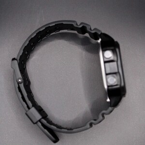 May include: A black and grey digital wristwatch with a rectangular face and a textured strap. The watch has three buttons on the side and a buckle fastening. The watch is designed for everyday use.