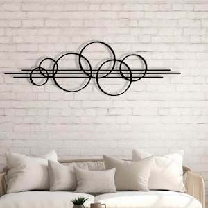 Puede incluir: Arte de pared de metal negro con círculos superpuestos y líneas horizontales. El diseño moderno está montado en una pared de ladrillo blanco sobre un sofá de color claro con cojines de tonos neutros. Una mesa de centro redonda de madera y plantas en macetas completan la escena.