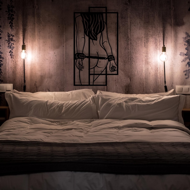 May include: A bedroom scene featuring a bed with white bedding and a dark gray blanket. Above the bed, a black metal wall art piece depicts a figure in a cage. Two wall-mounted light fixtures with exposed bulbs flank the art.