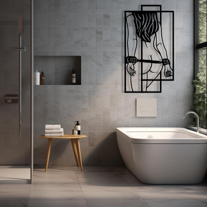 May include: Black metal wall art depicting a stylized female figure with long hair, arms behind the back, and cuffs. The artwork is mounted on a gray tiled wall in a bathroom setting. The bathroom includes a white bathtub, a small wooden table, and a shower.