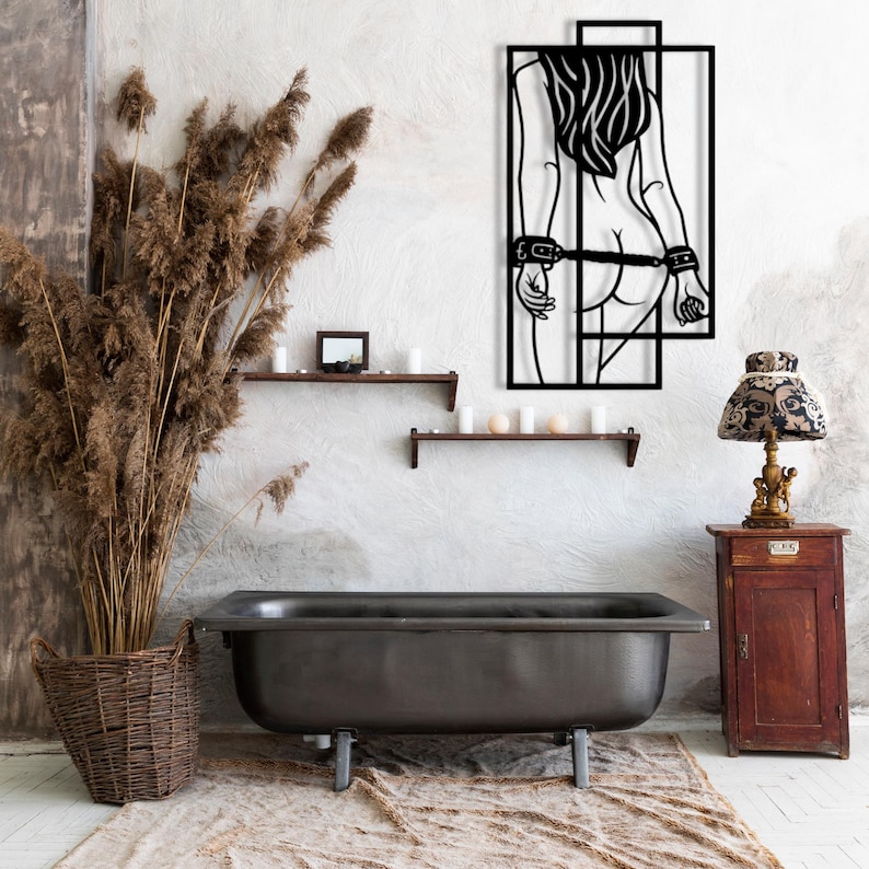 May include: Black metal wall art depicting a figure with long hair and handcuffs. The artwork is mounted on a textured white wall above a dark bathtub. The room is decorated with a lamp, shelves, and dried plants.