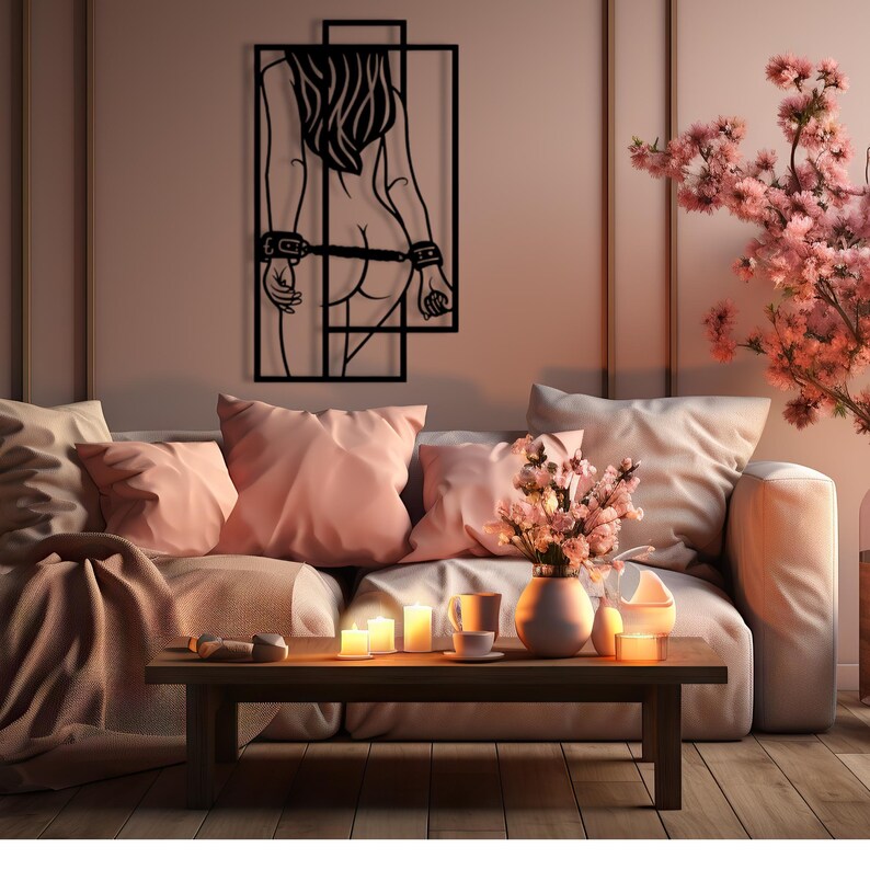 May include: A black metal wall art piece depicting a stylized female figure with her back to the viewer, framed by geometric lines. The figure has long hair and is bound at the wrists and waist. The artwork hangs above a sofa with pink pillows.