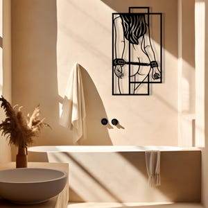 May include: Black metal wall art depicting a figure with long hair, hands cuffed behind the back, and a geometric frame. The artwork is mounted on a beige wall in a bathroom setting, with a bathtub and towel visible.