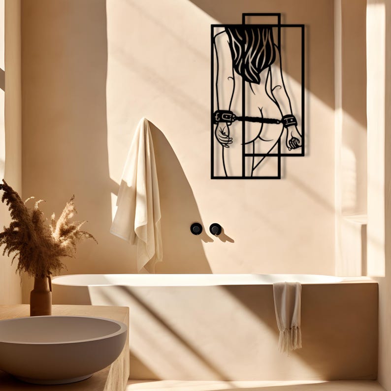 May include: Black metal wall art depicting a figure with long hair, hands cuffed behind the back, and a geometric frame. The artwork is mounted on a beige wall in a bathroom setting, with a bathtub and towel visible.