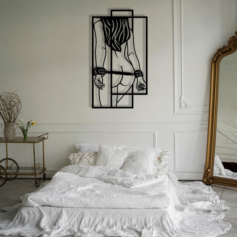 May include: Black metal wall art depicting a figure with long hair, hands cuffed behind the back, and a geometric frame. The artwork is mounted on a white wall above a bed with white linens. A gold-framed mirror is visible.