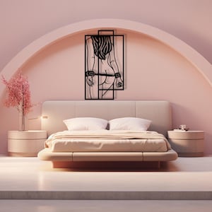 May include: A black metal wall art piece depicting a stylized female figure with handcuffs, framed within a rectangular design. The artwork is mounted on a pink wall above a bed with a beige headboard and bedding. The room has a modern, minimalist aesthetic.