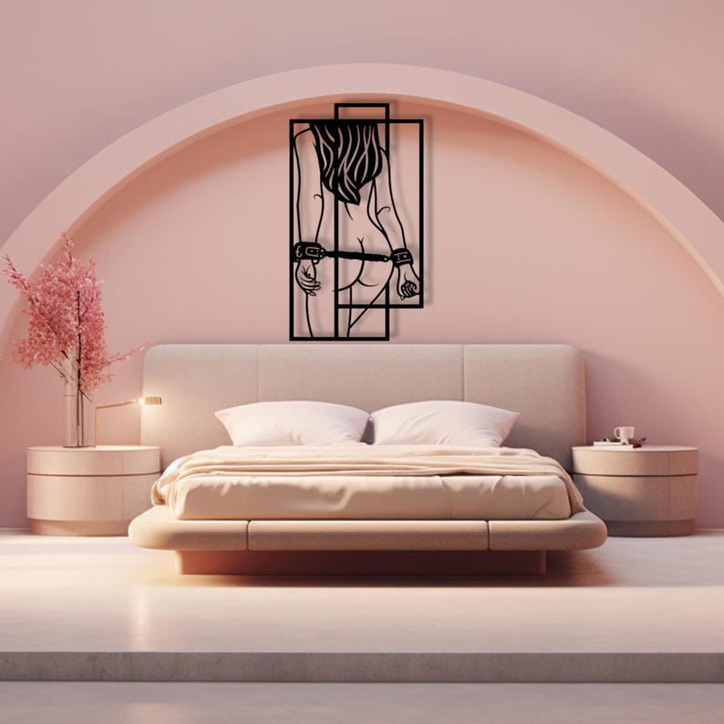 May include: A black metal wall art piece depicting a stylized female figure with handcuffs, framed within a rectangular design. The artwork is mounted on a pink wall above a bed with a beige headboard and bedding. The room has a modern, minimalist aesthetic.