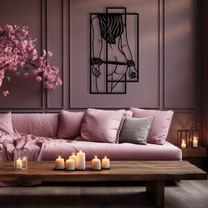 May include: A black metal wall art piece depicting a figure with their back to the viewer, bound at the wrists and waist. The art is mounted on a mauve wall above a pink sofa with pillows. A wooden coffee table holds lit candles.
