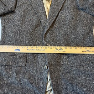 May include: A gray tweed blazer with a two-button closure and two front pockets. A wooden ruler is placed across the blazer, measuring its width. The blazer has a light-colored lining.