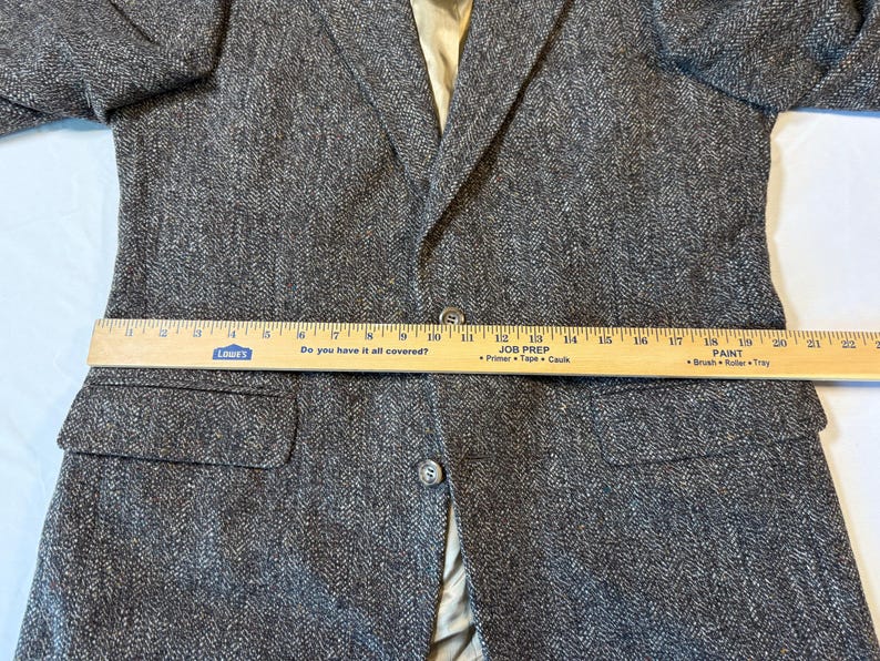 May include: A gray tweed blazer with a two-button closure and two front pockets. A wooden ruler is placed across the blazer, measuring its width. The blazer has a light-colored lining.