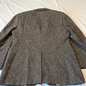 May include: Back view of a brown tweed blazer. The blazer has a notched lapel, two buttons, and a single vent in the back. The fabric has a textured appearance with flecks of brown and gray.
