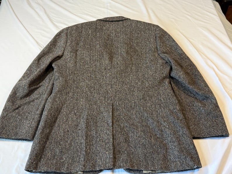 May include: Back view of a brown tweed blazer. The blazer has a notched lapel, two buttons, and a single vent in the back. The fabric has a textured appearance with flecks of brown and gray.