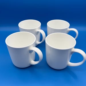 May include: Four white ceramic mugs with curved handles are arranged on a blue background. The mugs have a simple, clean design, suitable for coffee, tea, or other beverages. The mugs are evenly spaced.