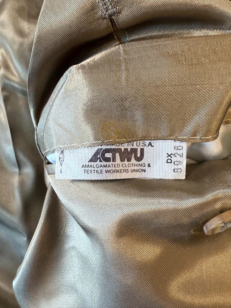 May include: Close-up of a tan garment with a tag that reads "MADE IN U.S.A." and "ACTWU AMALGAMATED CLOTHING & TEXTILE WORKERS UNION." The fabric has a subtle sheen.