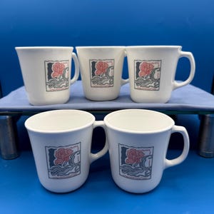 May include: Five white ceramic mugs with a curved handle. Each mug features a square design with a pink rose, gray waves, and a black border. The mugs are arranged on a blue surface.