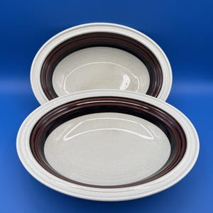 May include: Two oval-shaped serving bowls. The bowls have a speckled off-white interior and are bordered by a dark brown and black band. The rims are white. The bowls are set against a blue background.
