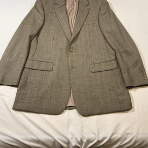 May include: A brown and tan houndstooth blazer with a two-button closure and two front pockets. The blazer has a notched lapel and a light brown interior lining. The sleeves have three buttons at the cuff. The blazer is laid flat on a white surface.
