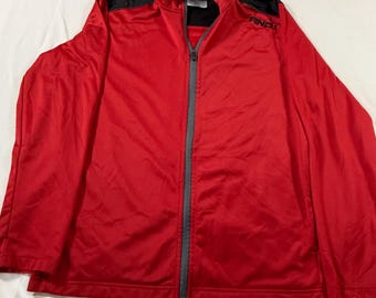 May include: A red athletic jacket with a black collar and shoulder accents. The jacket has a full-length gray zipper and the brand name "AND1" is visible on the right chest. The jacket is made of a smooth, shiny material.