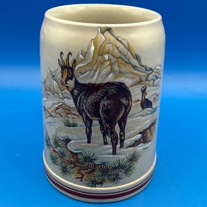 May include: A beige ceramic beer stein with a mountain and chamois design. The stein features a detailed illustration of a chamois in a snowy mountain landscape, with a red and white band around the base.