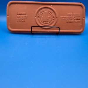 May include: A rectangular, brown bread basket brick with the text "BREAD BASKET BRICK" and "JOHANNABURG POTTERY" in a circular emblem. The text "USE WITH EXTREME CARE WHEN HOT" is also visible. A black metal stand supports the brick.