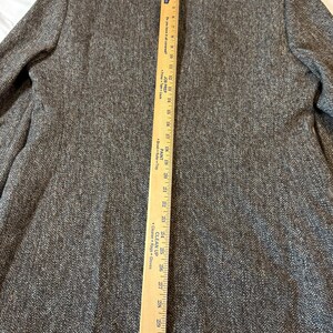 May include: A brown tweed blazer with a herringbone pattern. A wooden ruler is placed vertically along the back of the blazer, showing measurements in inches. The blazer has a notched lapel and two front pockets.
