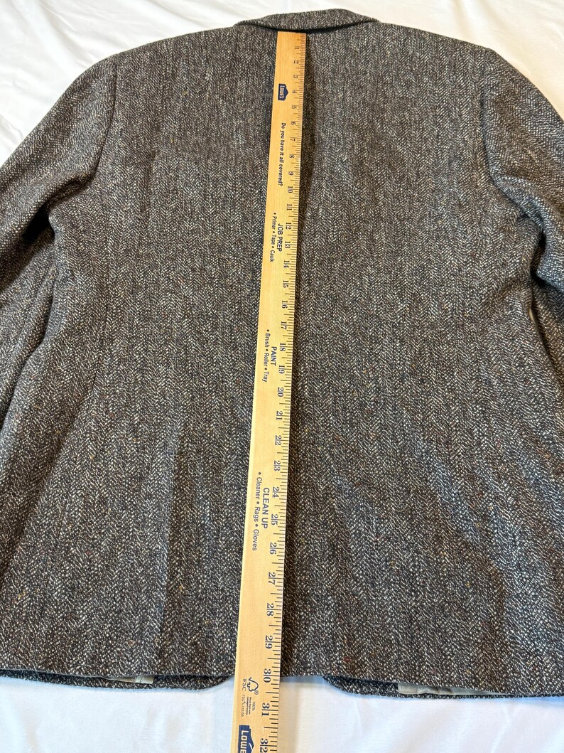 May include: A brown tweed blazer with a herringbone pattern. A wooden ruler is placed vertically along the back of the blazer, showing measurements in inches. The blazer has a notched lapel and two front pockets.