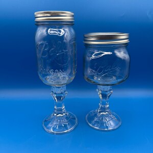 May include: Two clear glass goblets made from Ball Mason jars. The jars have silver lids and are mounted on decorative glass stems. The jars are embossed with the Ball logo and the word "MASON".