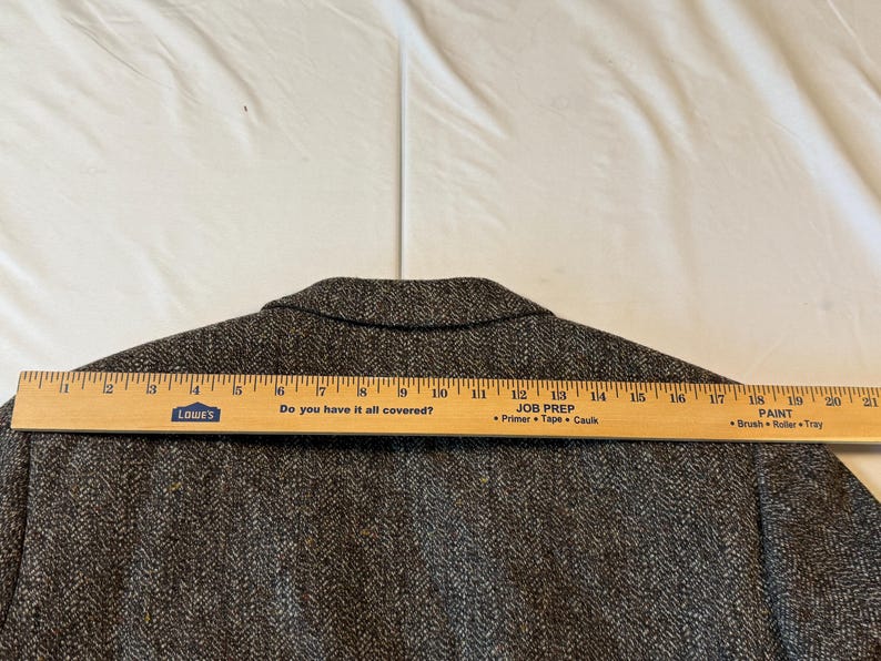 May include: A brown tweed blazer with a wooden ruler laid across the back. The ruler measures approximately 20 inches. The blazer has a herringbone pattern and a notched collar. The text "Do you have it all covered?" is visible on the ruler.
