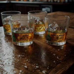 May include: Four glasses of Usher's Green Stripe Scotch Whisky on a wooden surface. Each glass contains amber-colored liquid and ice cubes. The Usher's logo and text are visible on the glasses, with the words "Scotch Whisky" and "Bottled in Scotland."