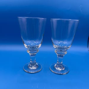 May include: Two clear glass goblets with a smooth, rounded bowl shape, a short, thick stem featuring a decorative ring detail, and a wide, circular base. The glassware is transparent and features a simple, functional design for beverages.
