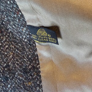 May include: Close-up of a tweed fabric with a dark brown and white speckled pattern. A black tag with gold text reads "100% PURE WOOL DRY CLEAN ONLY". The lining is a light brown color.
