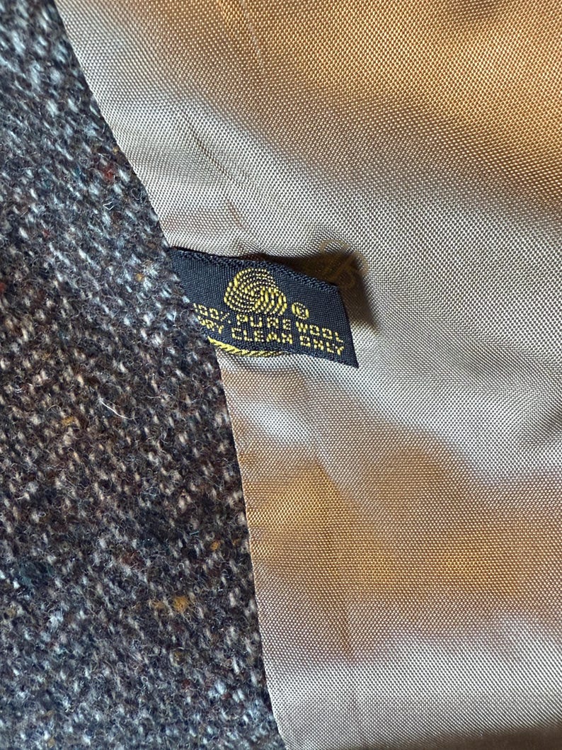 May include: Close-up of a tweed fabric with a dark brown and white speckled pattern. A black tag with gold text reads "100% PURE WOOL DRY CLEAN ONLY". The lining is a light brown color.