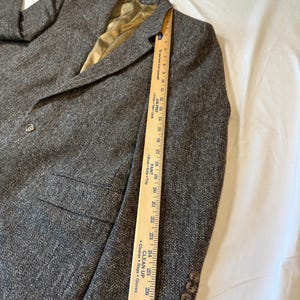May include: A gray tweed blazer with a notched lapel, two front pockets, and three buttons on the cuff. The blazer has a single-button closure and a gold-colored lining. A wooden ruler is placed along the side of the blazer.