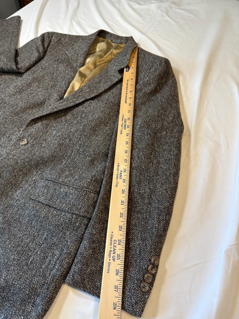 May include: A gray tweed blazer with a notched lapel, two front pockets, and three buttons on the cuff. The blazer has a single-button closure and a gold-colored lining. A wooden ruler is placed along the side of the blazer.