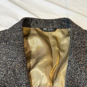 May include: Close-up of a tweed blazer with a textured, speckled pattern in shades of gray, brown, and black. The interior lining is a golden-tan color. The label reads "BILL BLASS".