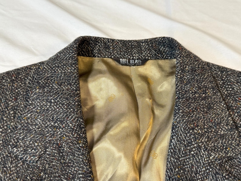 May include: Close-up of a tweed blazer with a textured, speckled pattern in shades of gray, brown, and black. The interior lining is a golden-tan color. The label reads "BILL BLASS".