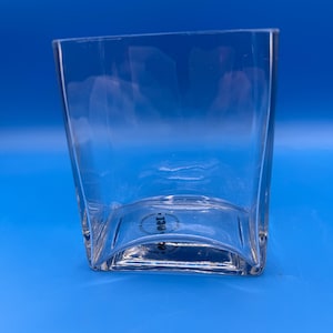 May include: A clear, square-shaped glass with a curved base. The glass has a slightly tapered design, wider at the top than the base. The glass is empty and sits against a solid blue background.