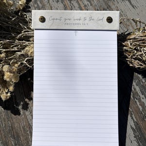 May include: A white lined notepad with a marble-patterned top header secured by two bronze-colored screws. The header features the text 'Commit your work to the Lord' and 'PROVERBS 16:3' with a small cross symbol printed above the lines.