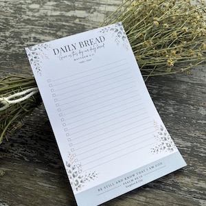 May include: A white rectangular notepad featuring a checklist with horizontal lines. The header reads DAILY BREAD Give us this day our daily bread MATTHEW 6:11. The footer reads BE STILL AND KNOW THAT I AM GOD PSALM 46:10 Kingdom & Clay.