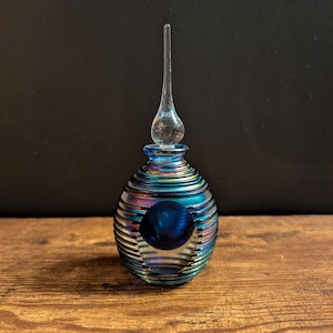 Puede incluir: Un pequeño frasco de perfume de vidrio iridiscente con un tapón largo y delgado. El frasco tiene un cuerpo esférico con estrías horizontales y un orbe azul oscuro visible a través del centro. Los colores cambian de azul a verde y morado.
